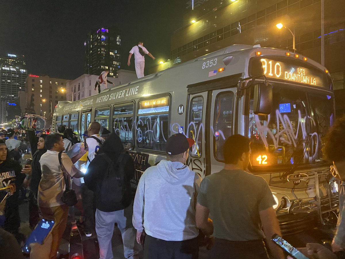 There are a dozen people on top of a graffitied city bus with a RAMS HOUSE flag and a detour sign and street cones. The bus driver is inside. Police in copter above. Fireworks constantly going off