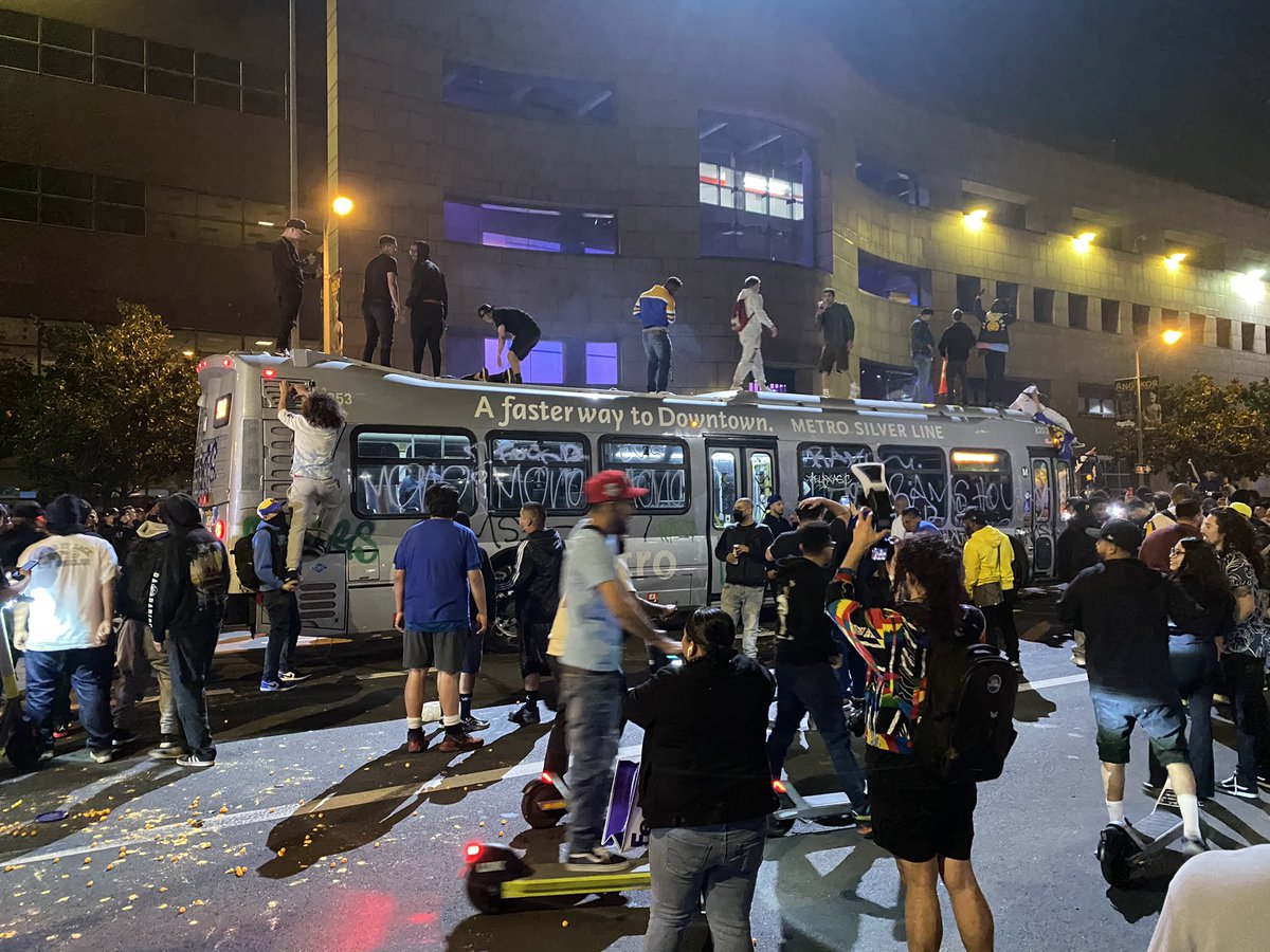 There are a dozen people on top of a graffitied city bus with a RAMS HOUSE flag and a detour sign and street cones. The bus driver is inside. Police in copter above. Fireworks constantly going off