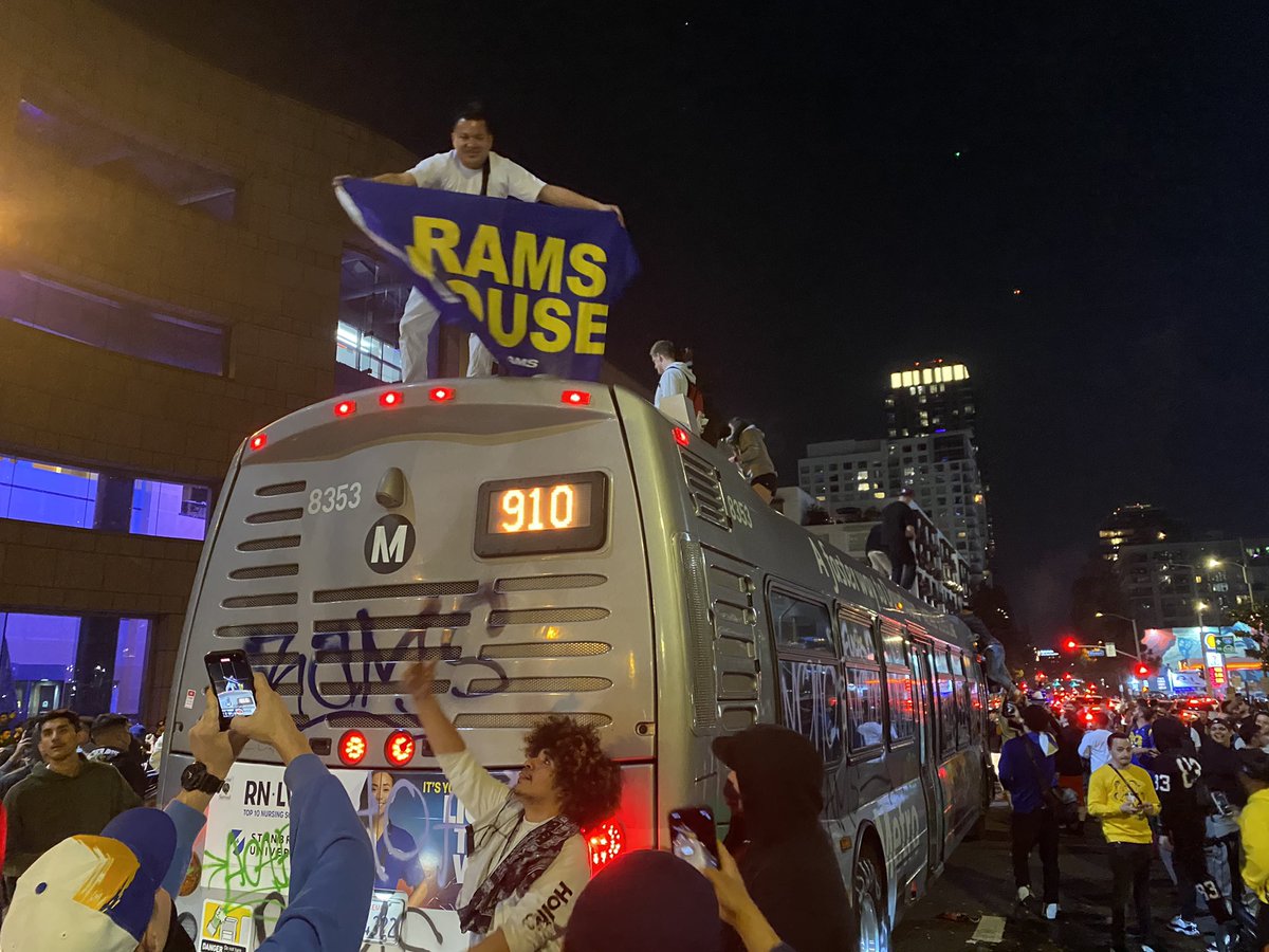 There are a dozen people on top of a graffitied city bus with a RAMS HOUSE flag and a detour sign and street cones. The bus driver is inside. Police in copter above. Fireworks constantly going off